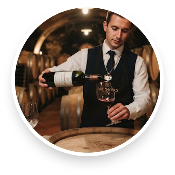 Un sommelier versant du vin rouge dans un verre dans une cave à vin bordée de tonneaux en bois.